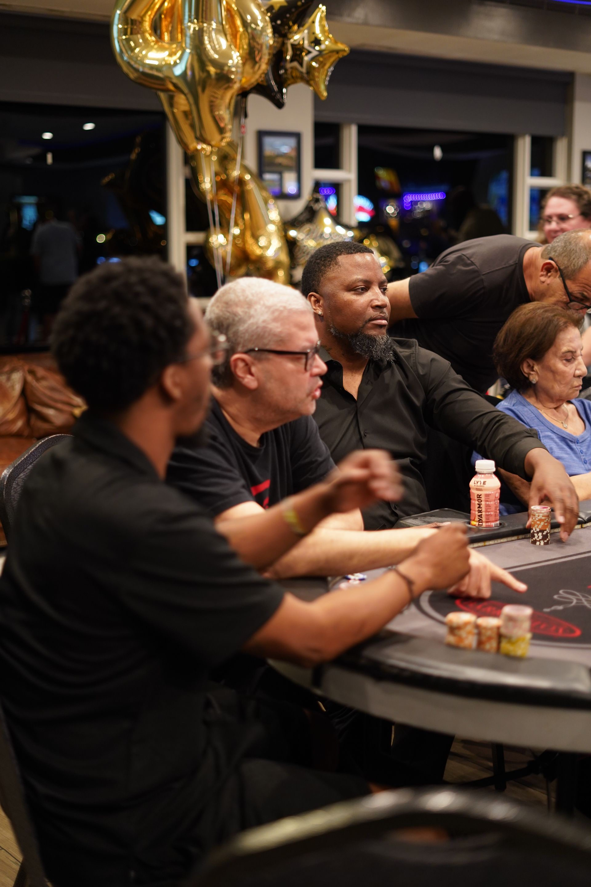 People seated at a poker table in a casino, with gold balloons and chips visible in the foreground.