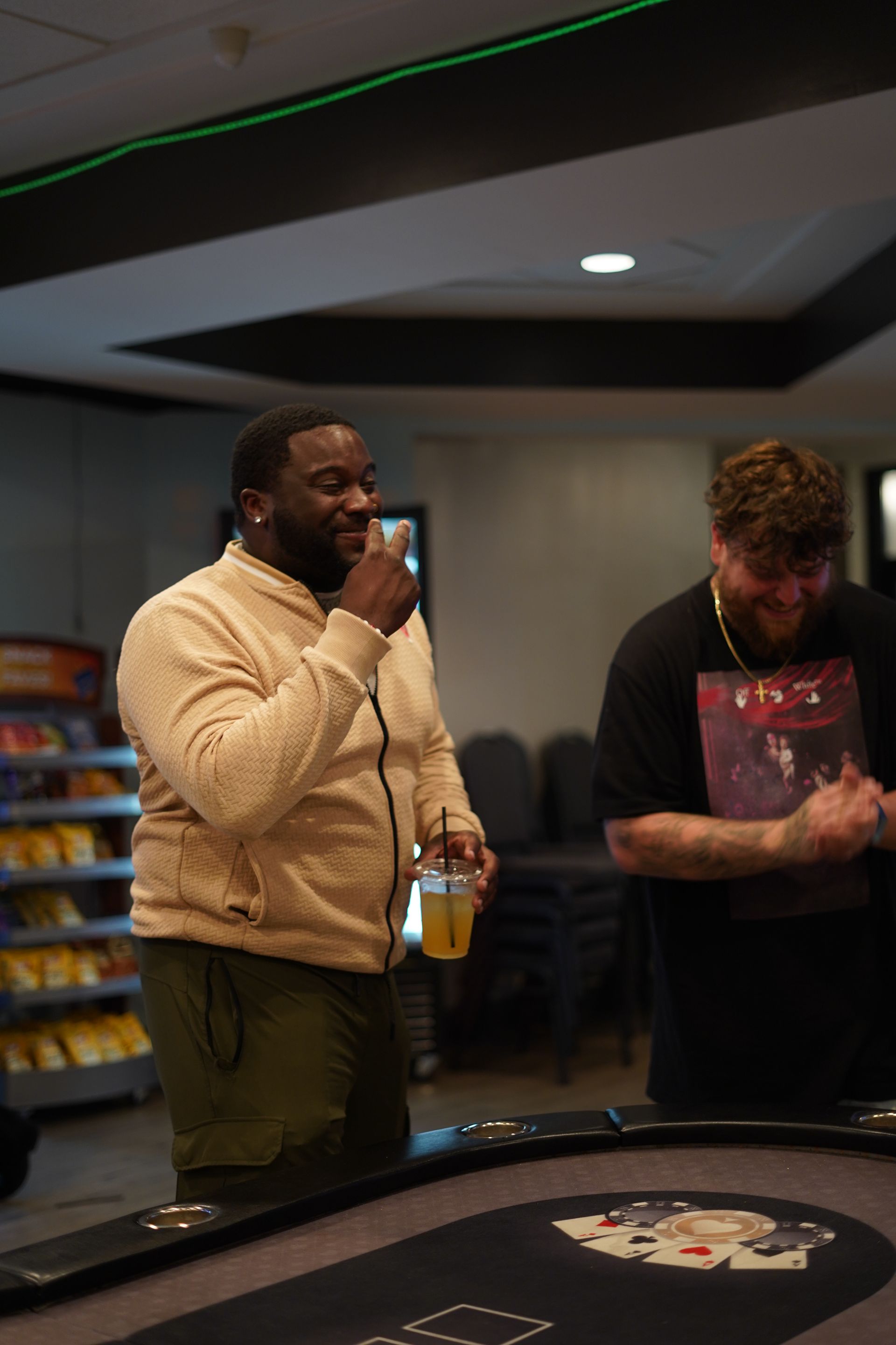 Two men standing at a casino table, one drinking from a cup and the other looking down at his hands.