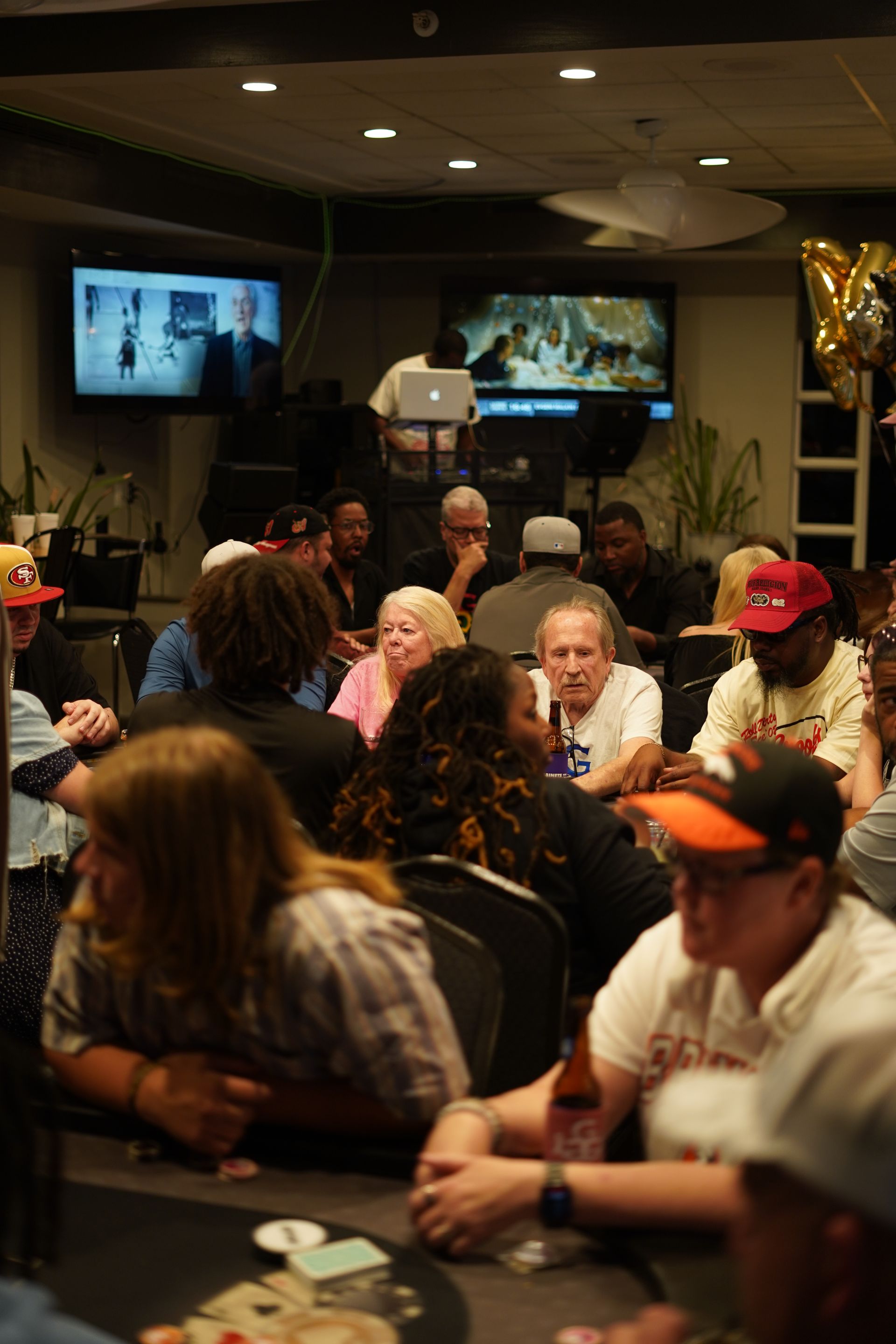 Crowded indoor gathering with people seated at tables and two wall-mounted screens in the background
