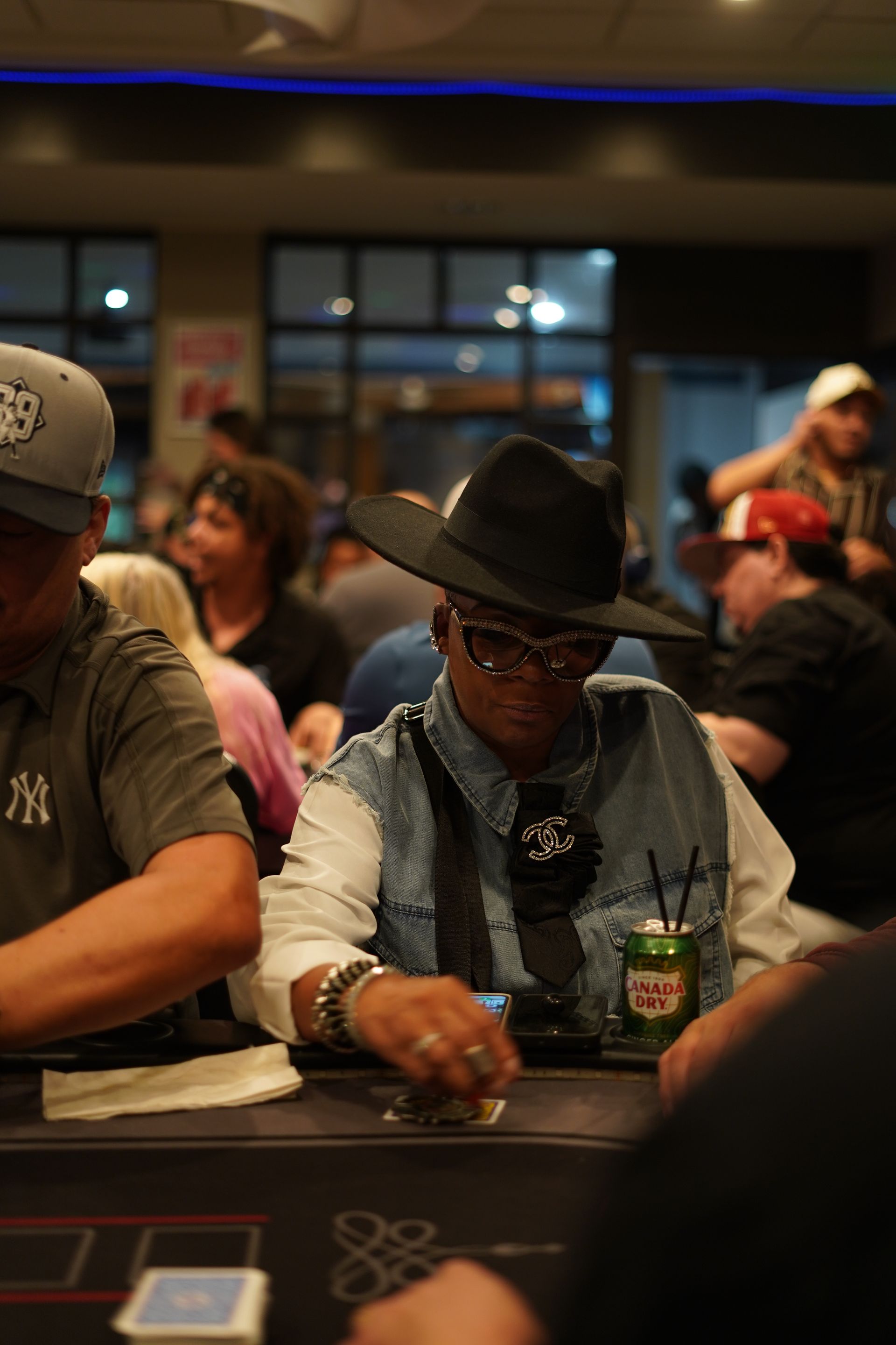 Man in black hat seated at a casino table with a drink, surrounded by other players in a dim room