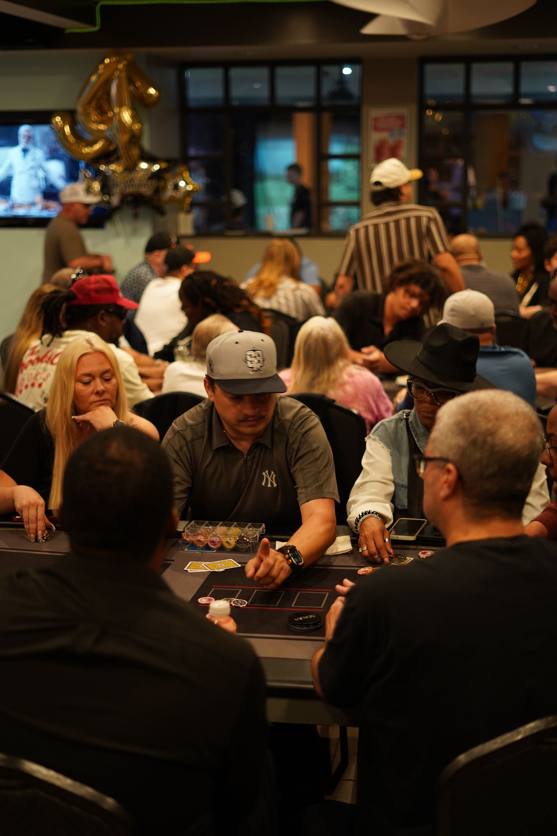 People seated at tables in a casino or card room, with gold balloon decorations in the background.