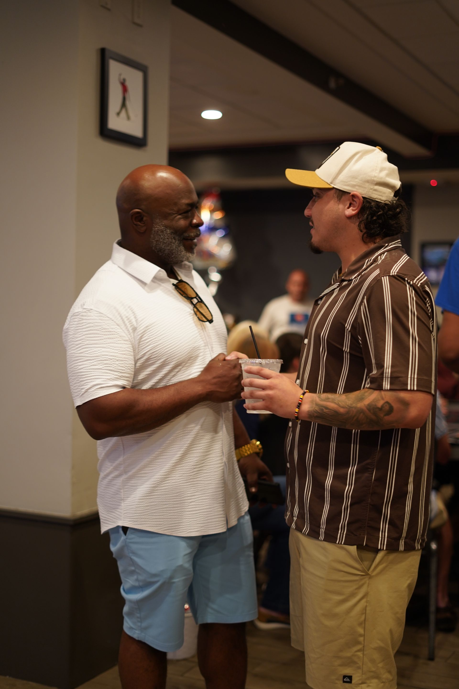 Two men talking in a hallway, one holding a drink, with event posters on the wall.