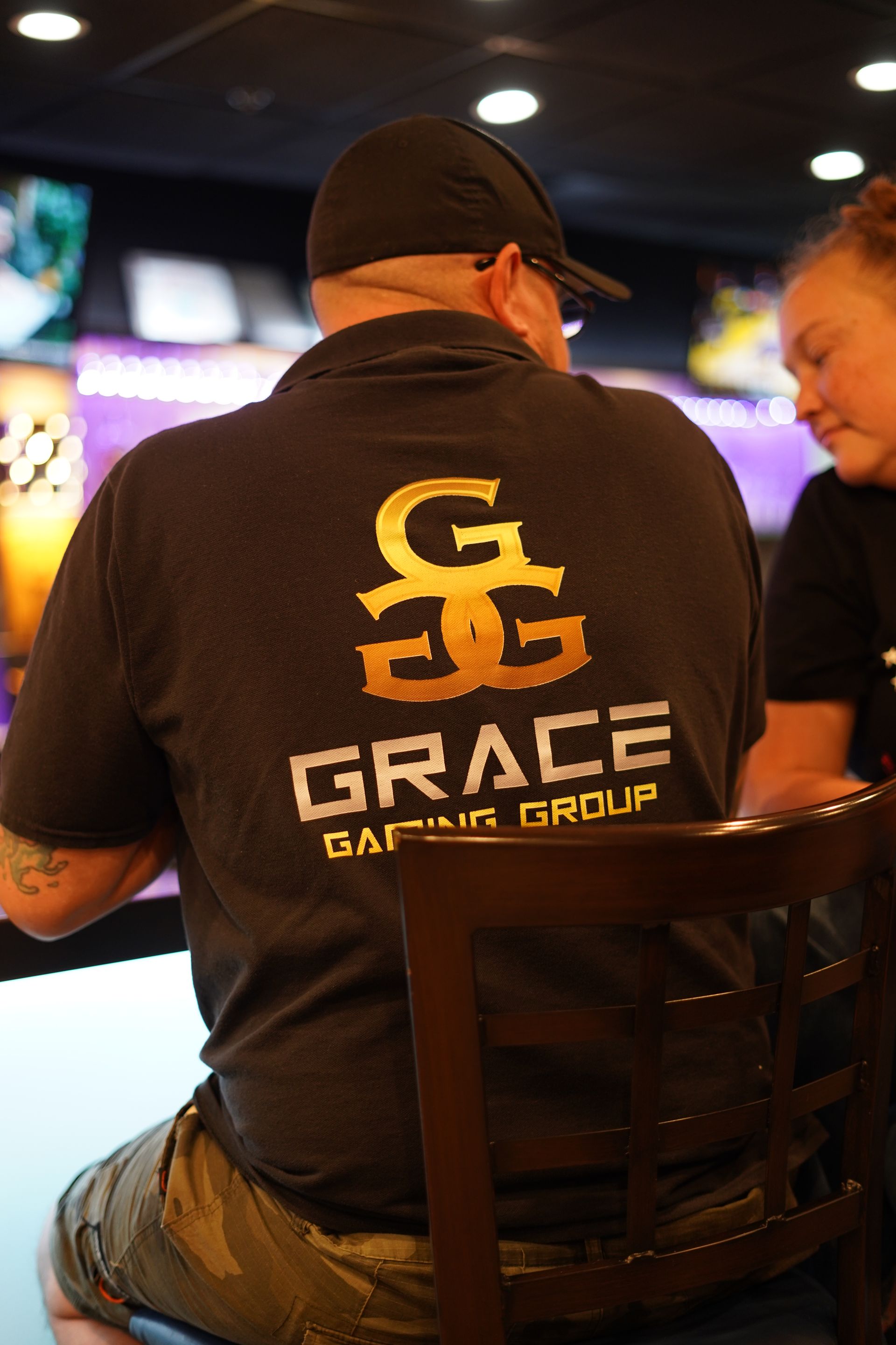 Person in black cap and Grace Gaming Group shirt seated in a dimly lit gaming venue