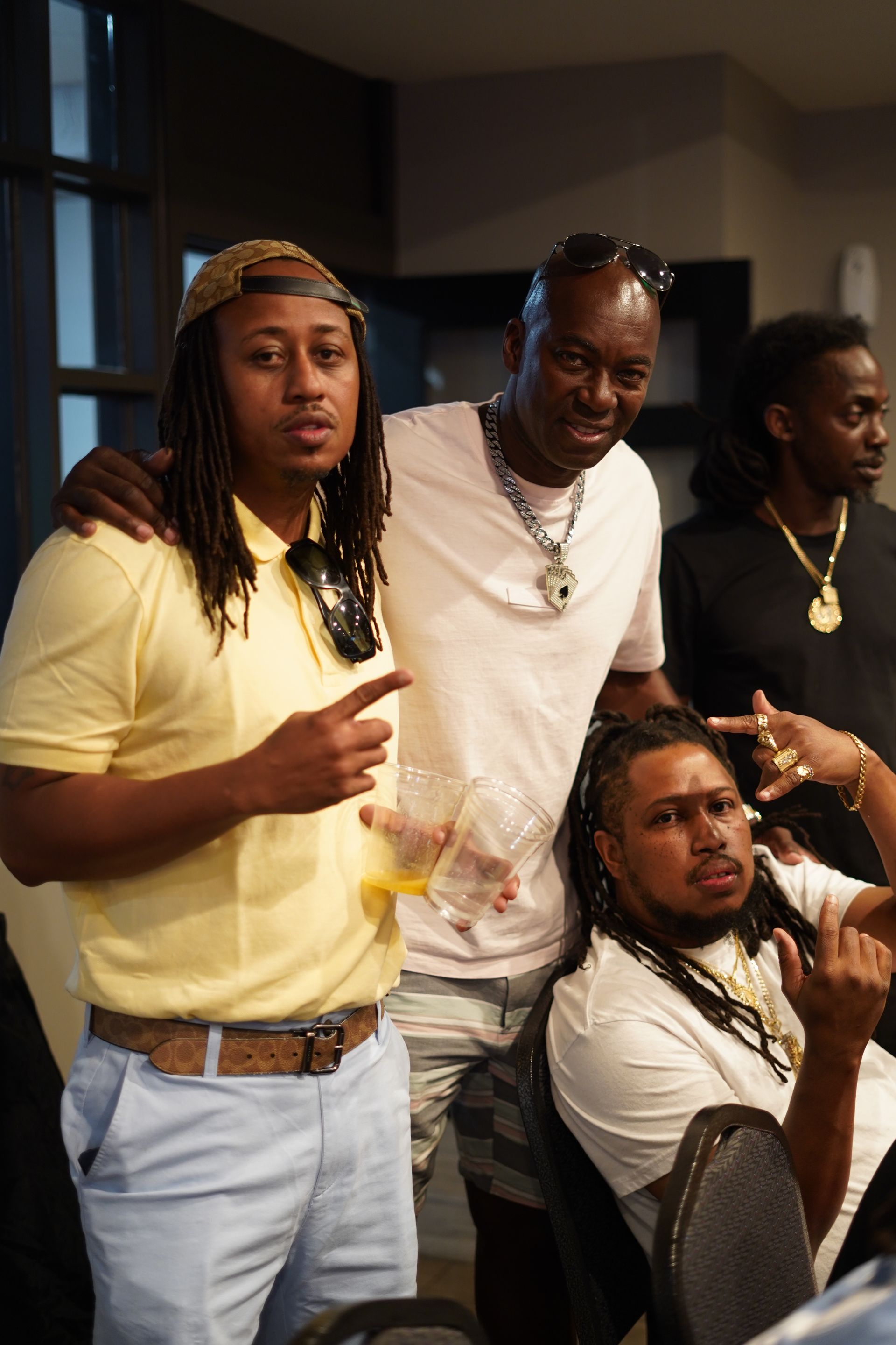 Four people posing indoors, two standing and one seated, wearing casual clothes and gold chains.