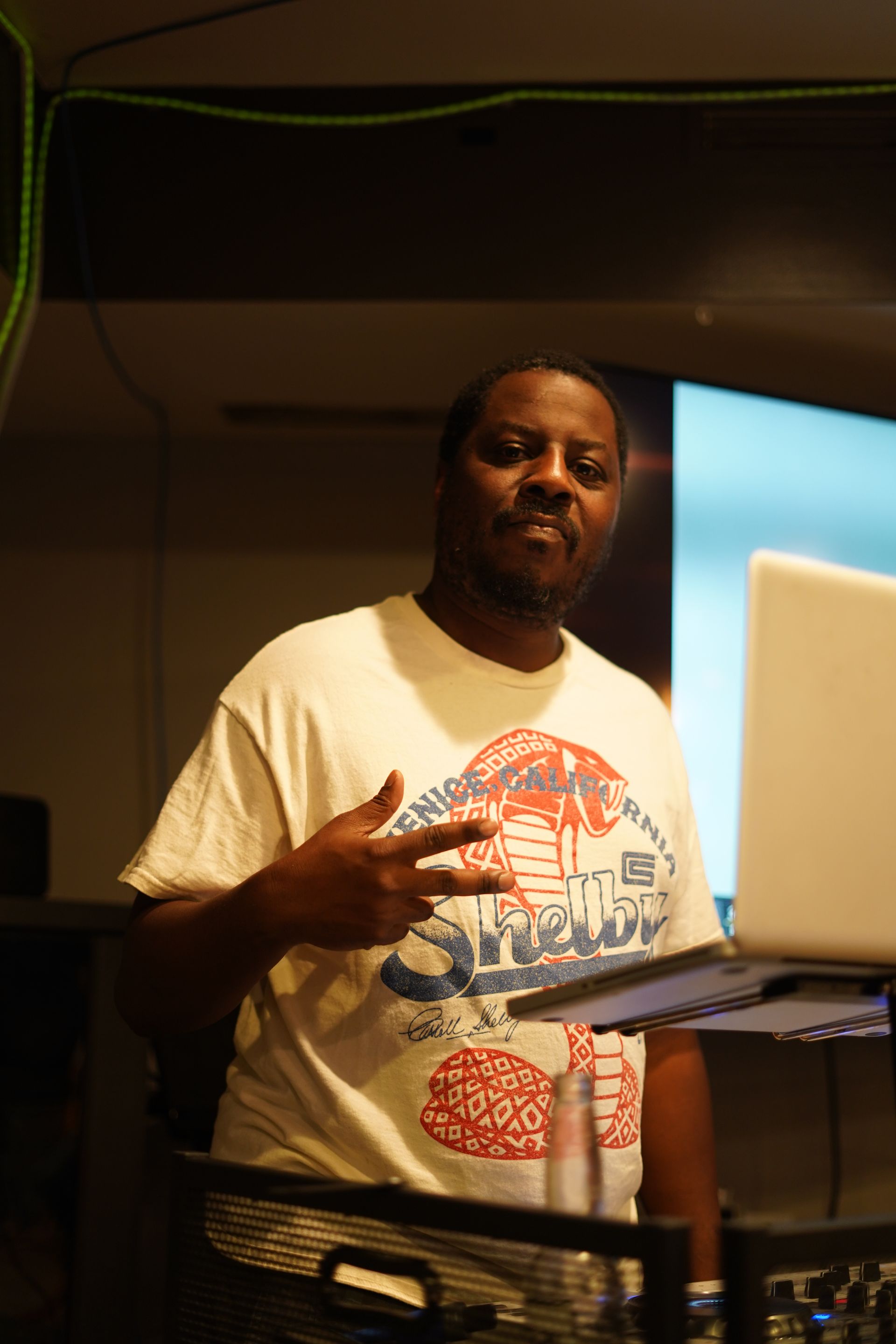 Man standing beside a laptop in a dim room, making a hand gesture near a blue-lit screen