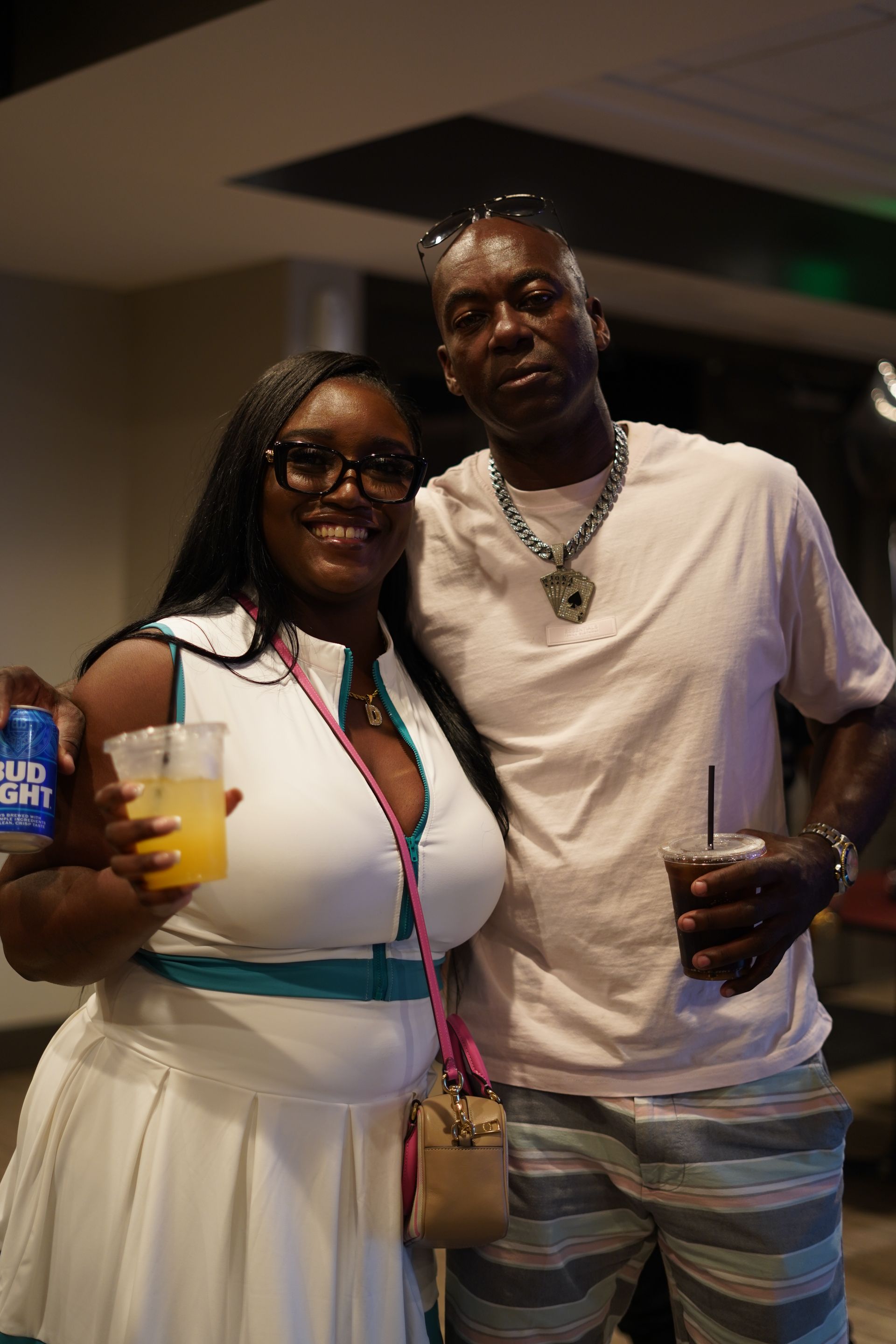 Two people pose indoors, smiling and holding drinks, dressed in white and pastel summer outfits.