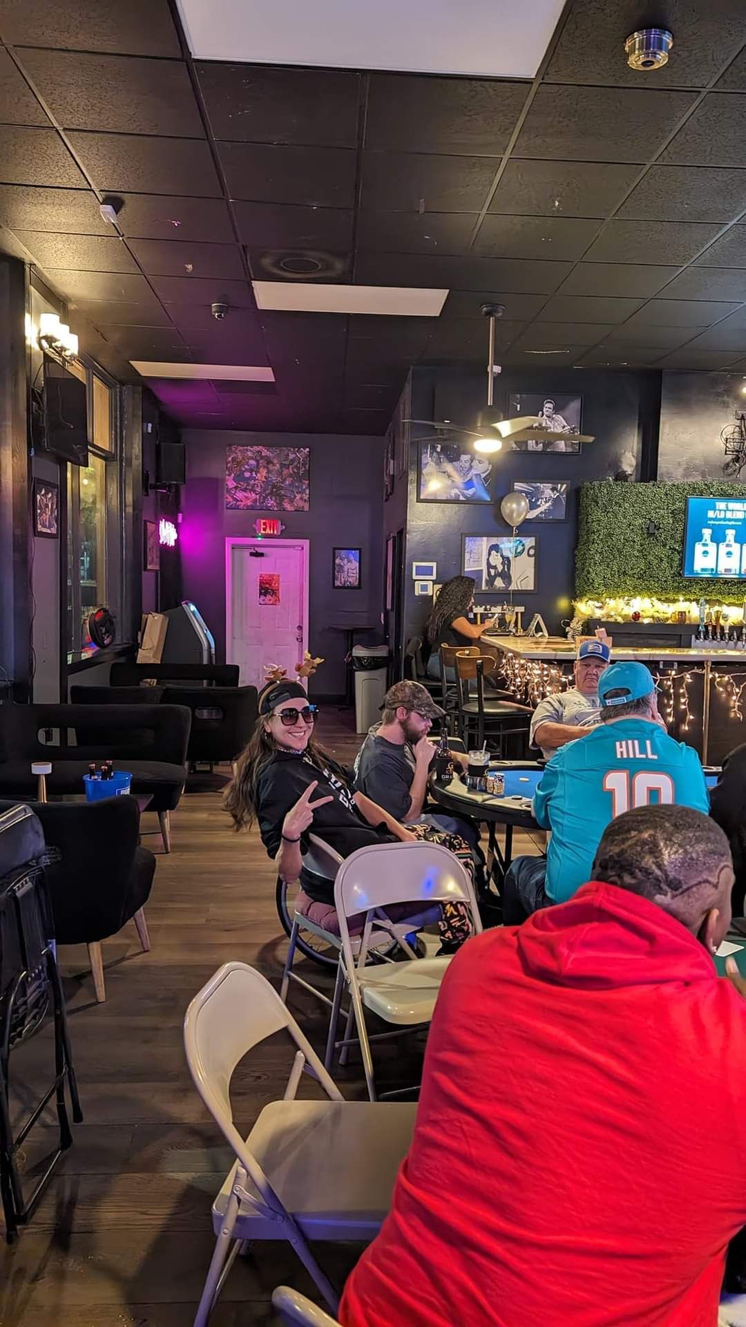 People seated at tables in a dim bar with a lit TV screen and neon purple lighting.