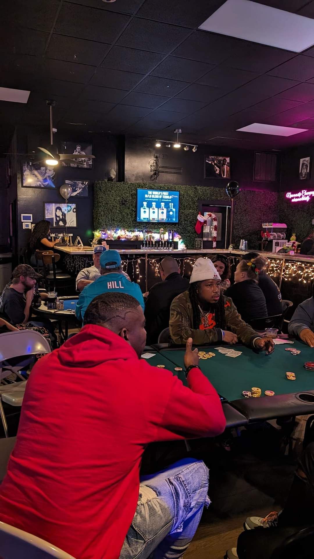People playing pool in a dimly lit bar, with a neon TV screen and red and green jackets in the foreground