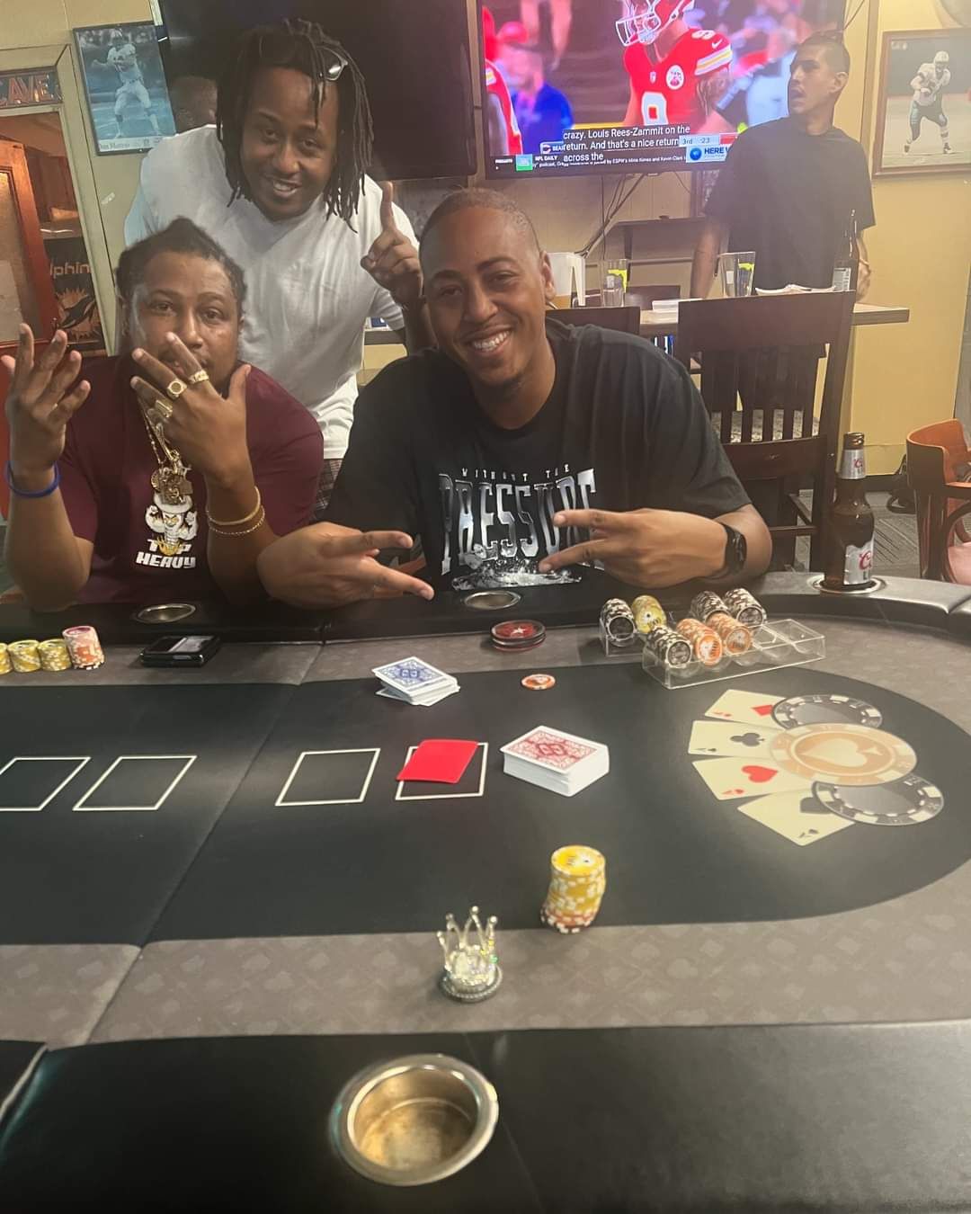 Three people smiling at a casino table with cards, chips, and slot machines in the background