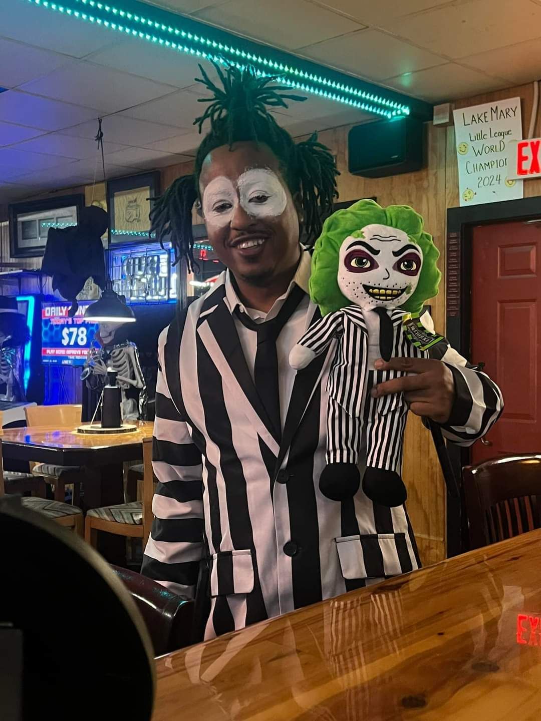 Person in a clown outfit holding a doll at a bar, with neon lights and an EXIT sign in the background.