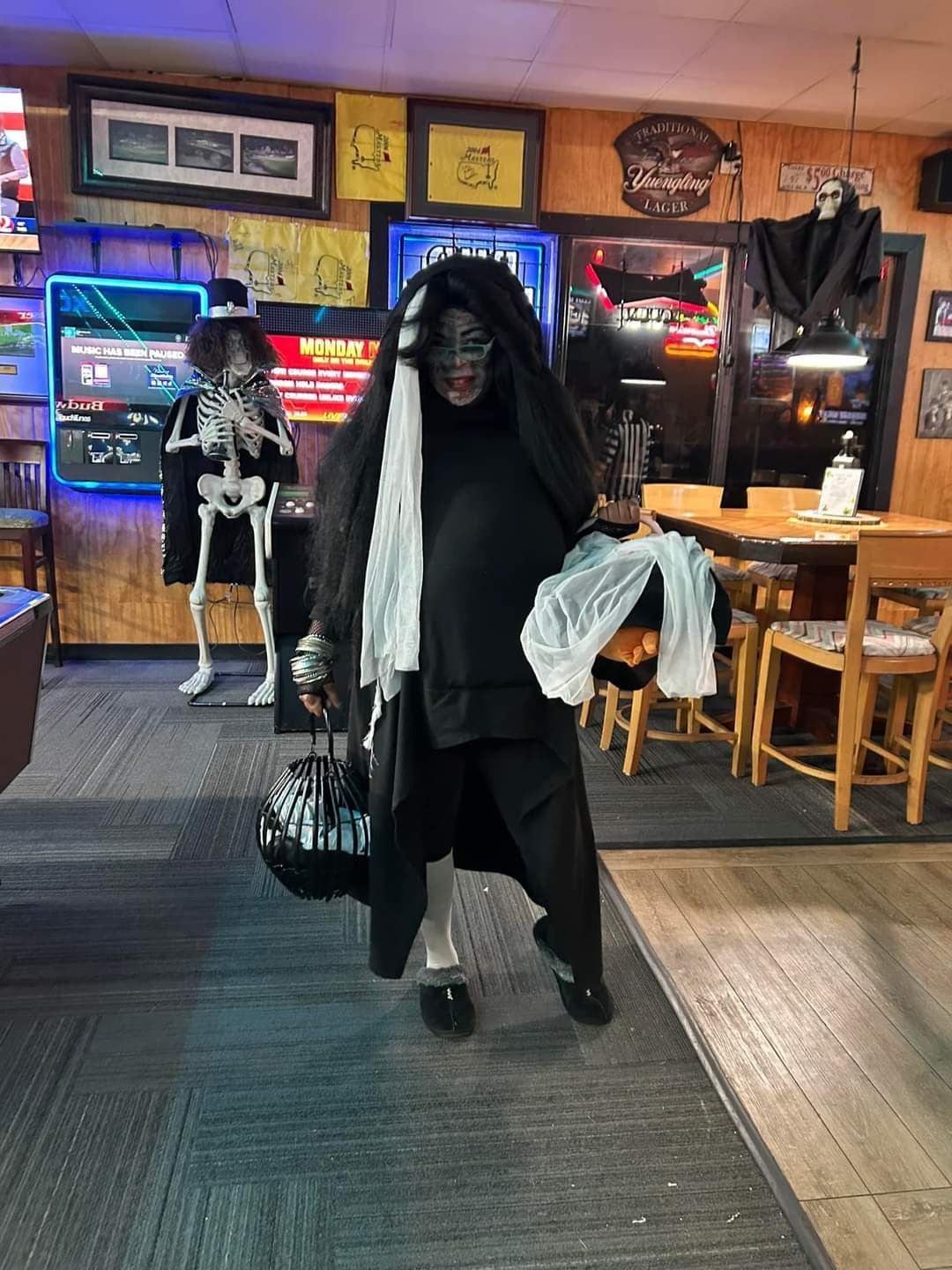 Halloween-costumed person in black cloak and mask standing in a dimly lit bar, holding a bag and draped fabric