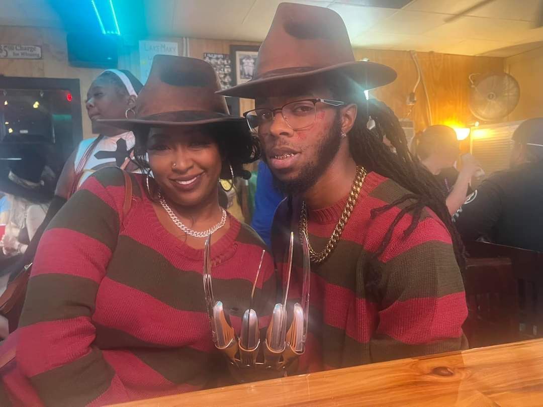 Two people in matching striped sweaters and hats pose at a party, smiling under warm lights.