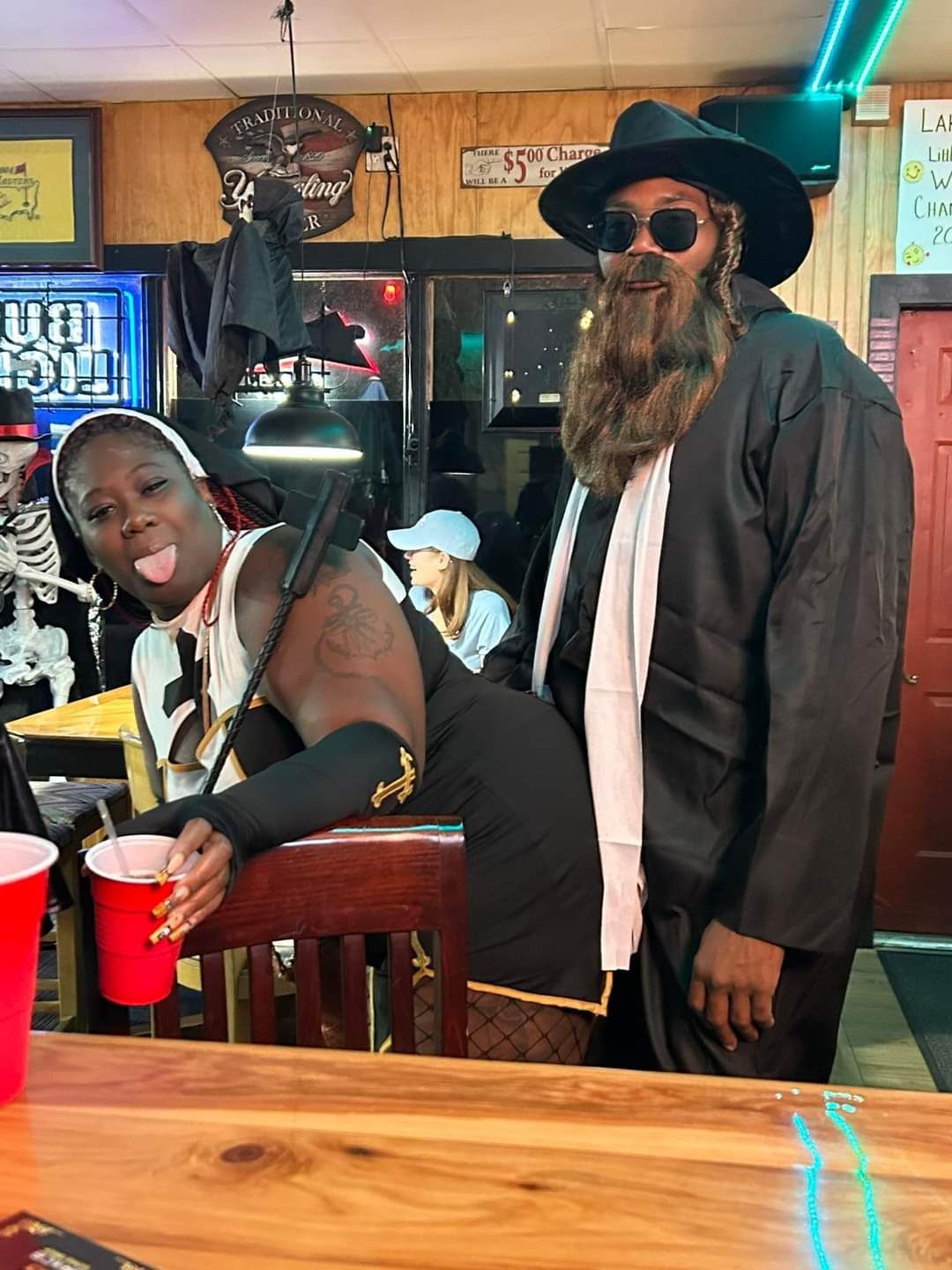 Two people posing at a bar, one leaning over with red cups on a wooden table.