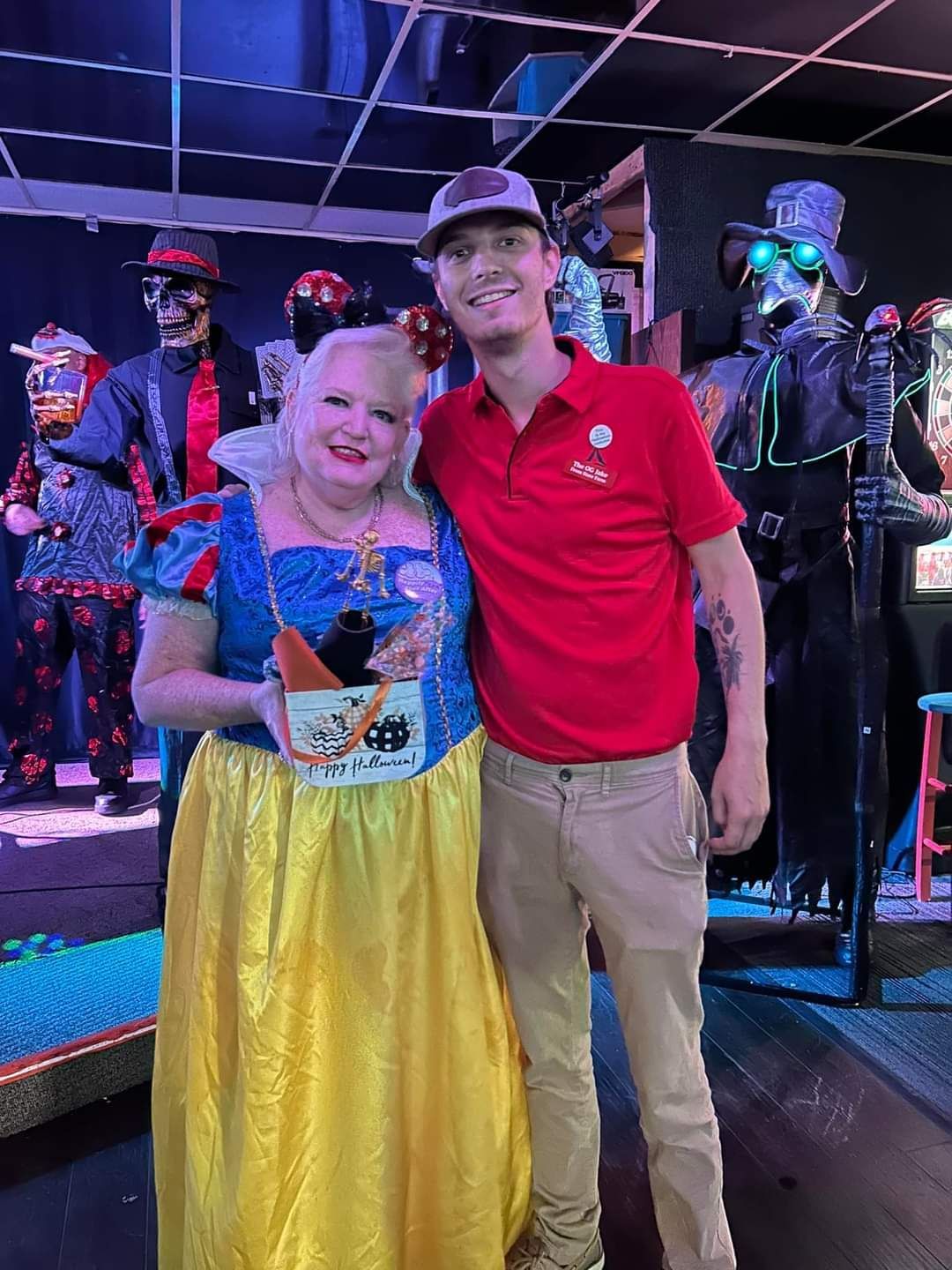 Two smiling people pose in colorful outfits at a neon-lit event with costumed performers in the background