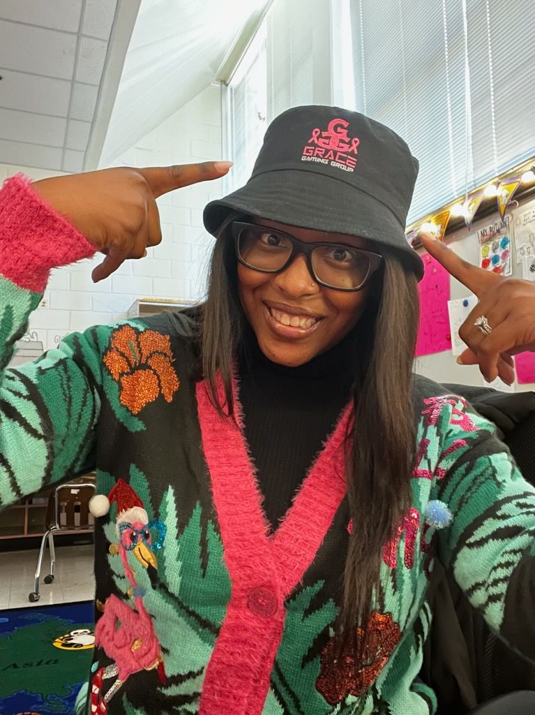 Smiling person in glasses and black cap points at the cap, wearing a colorful patterned sweater.