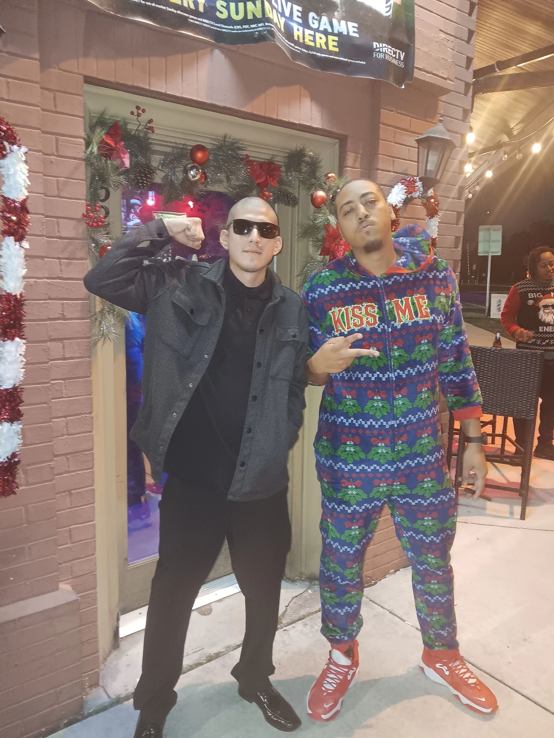 Two people posing outside a storefront, one in black and one in a colorful sweater and red sneakers.