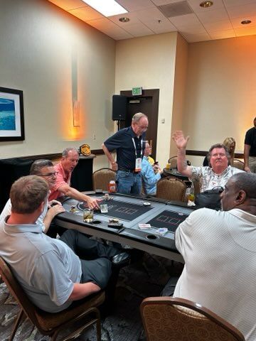 People gathered around a card table in a hotel meeting room, one standing and raising a hand.