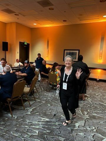 Woman waving in a hotel banquet hall with seated guests and warm orange lighting.