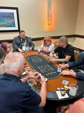 People seated around a casino table, holding cards and chips in a warmly lit room.