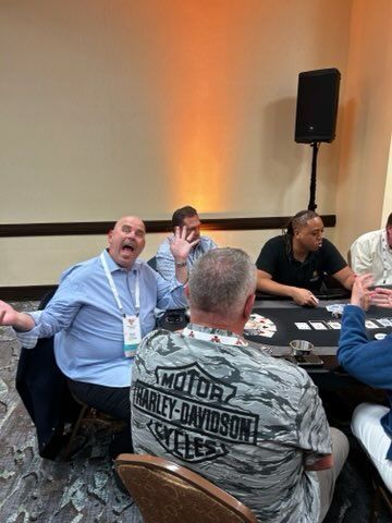 Men seated at round tables in a conference room, one man laughing near a Harley-Davidson shirt.
