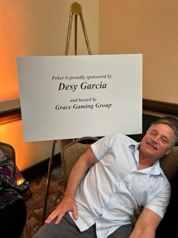Man reclining beside a white sign on a gold easel, smiling in a warmly lit room