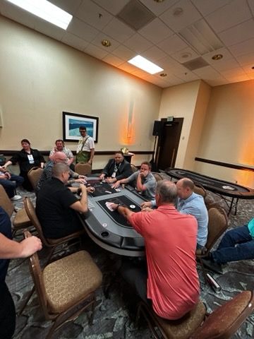 People seated around a casino poker table in a carpeted conference room, playing cards and chips.