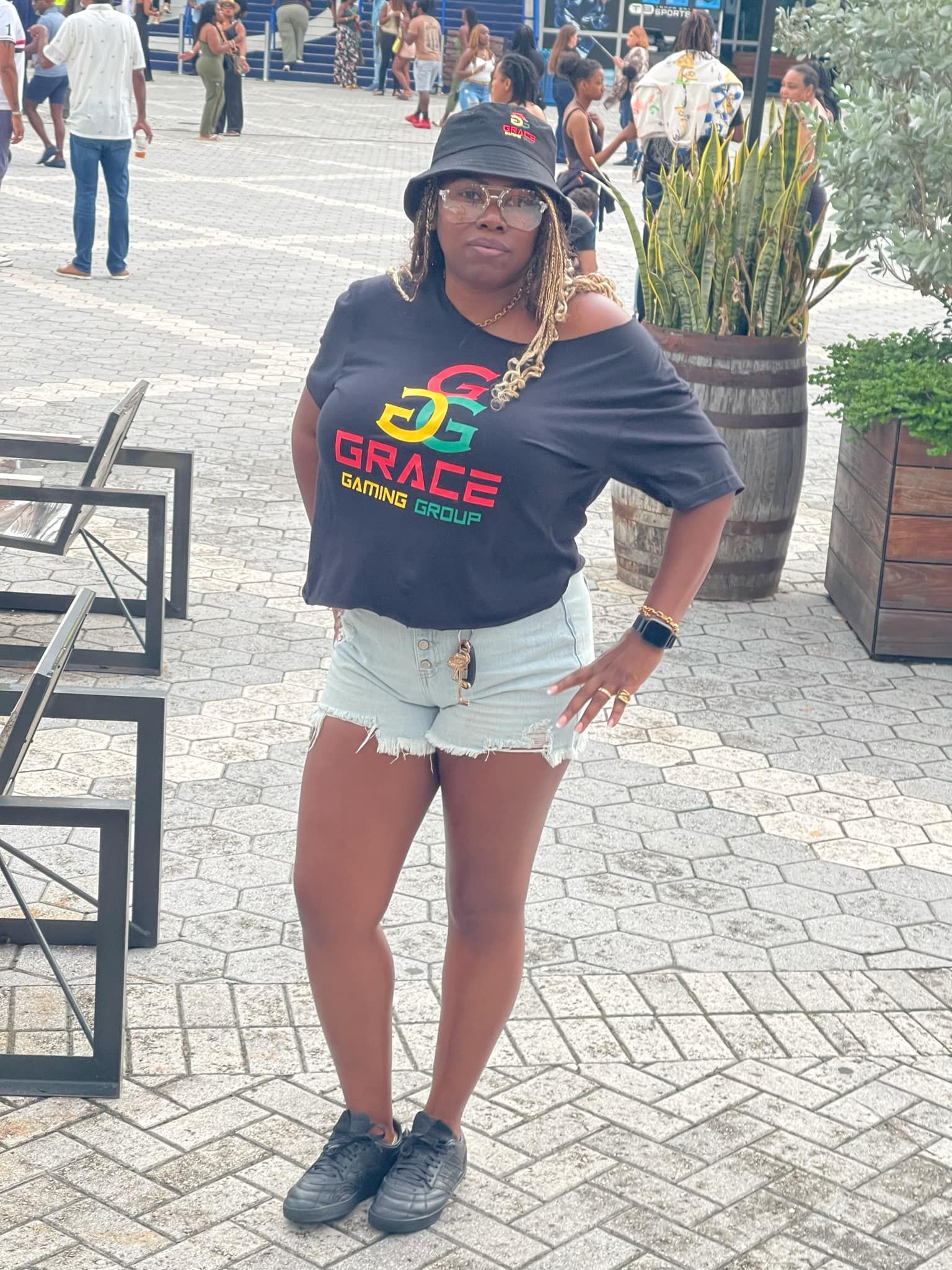 Person posing outdoors in a black “GRACE” T-shirt, denim shorts, and hat on a brick walkway.