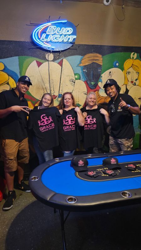 Five people posing by a casino table under a blue neon sign, wearing black T-shirts and smiling.