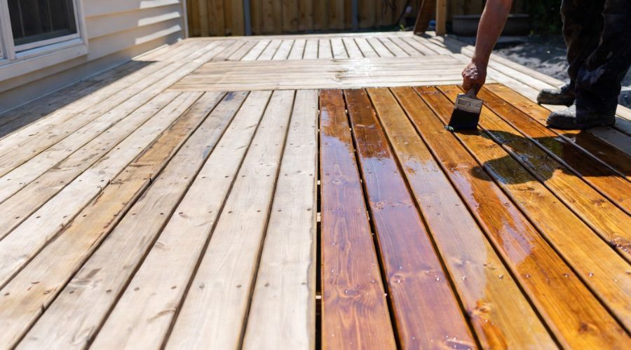 A person brushes a warm, glossy stain onto one half of a weathered wooden deck, contrasting it with the dry, grayed wood.