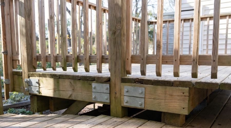 A low-angle view of a wooden deck frame, showing bolted metal support plates attaching horizontal beams to support posts.