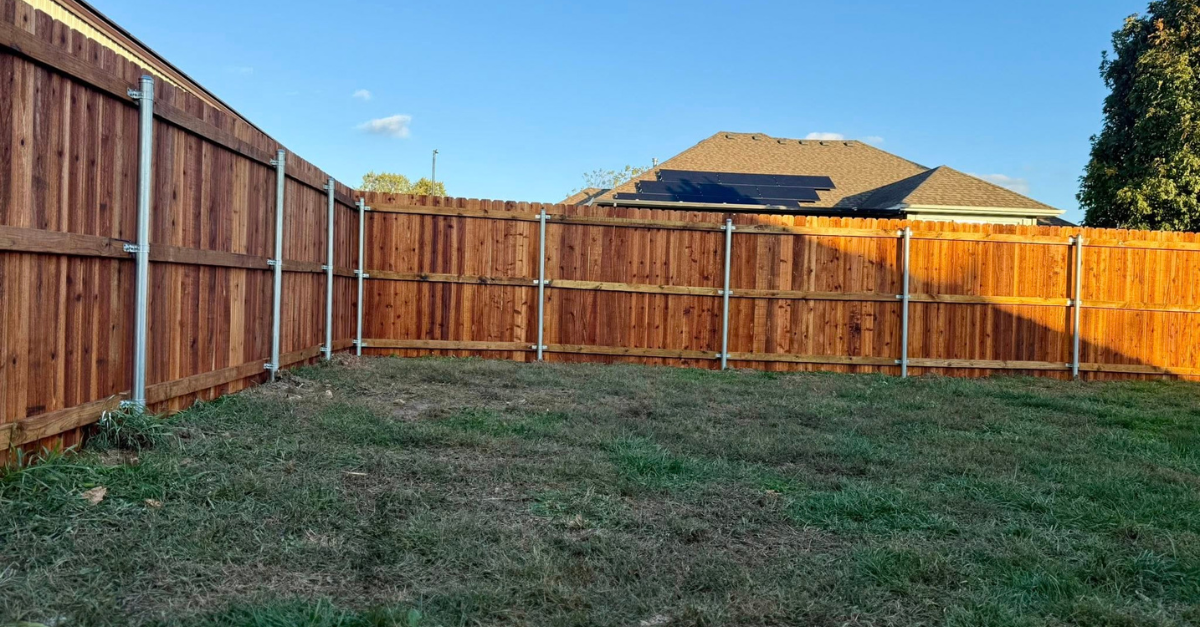 Wooden fence surrounding a grassy backyard with a house in the background. Bright blue sky.