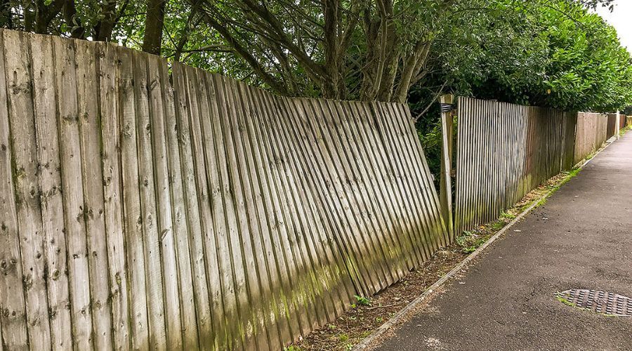 Leaning wooden privacy fence damaged by a severe storm alongside a paved neighborhood sidewalk.