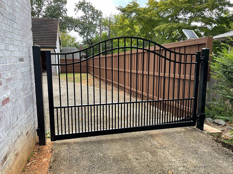 A black metal gate is open to a driveway next to a wooden fence.
