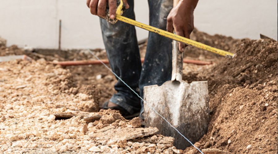 Worker measuring ground depth to dig a fence post hole in rocky soil.