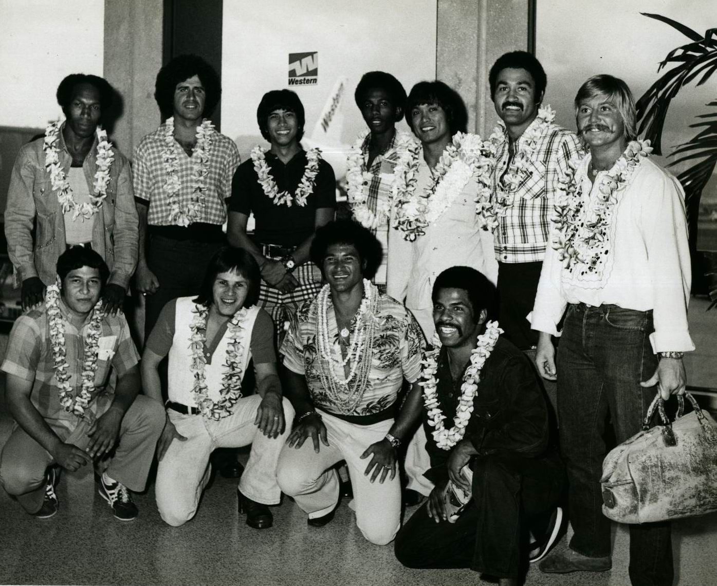 Group of people wearing leis, likely arriving at an airport. Most are smiling, dressed in casual attire.