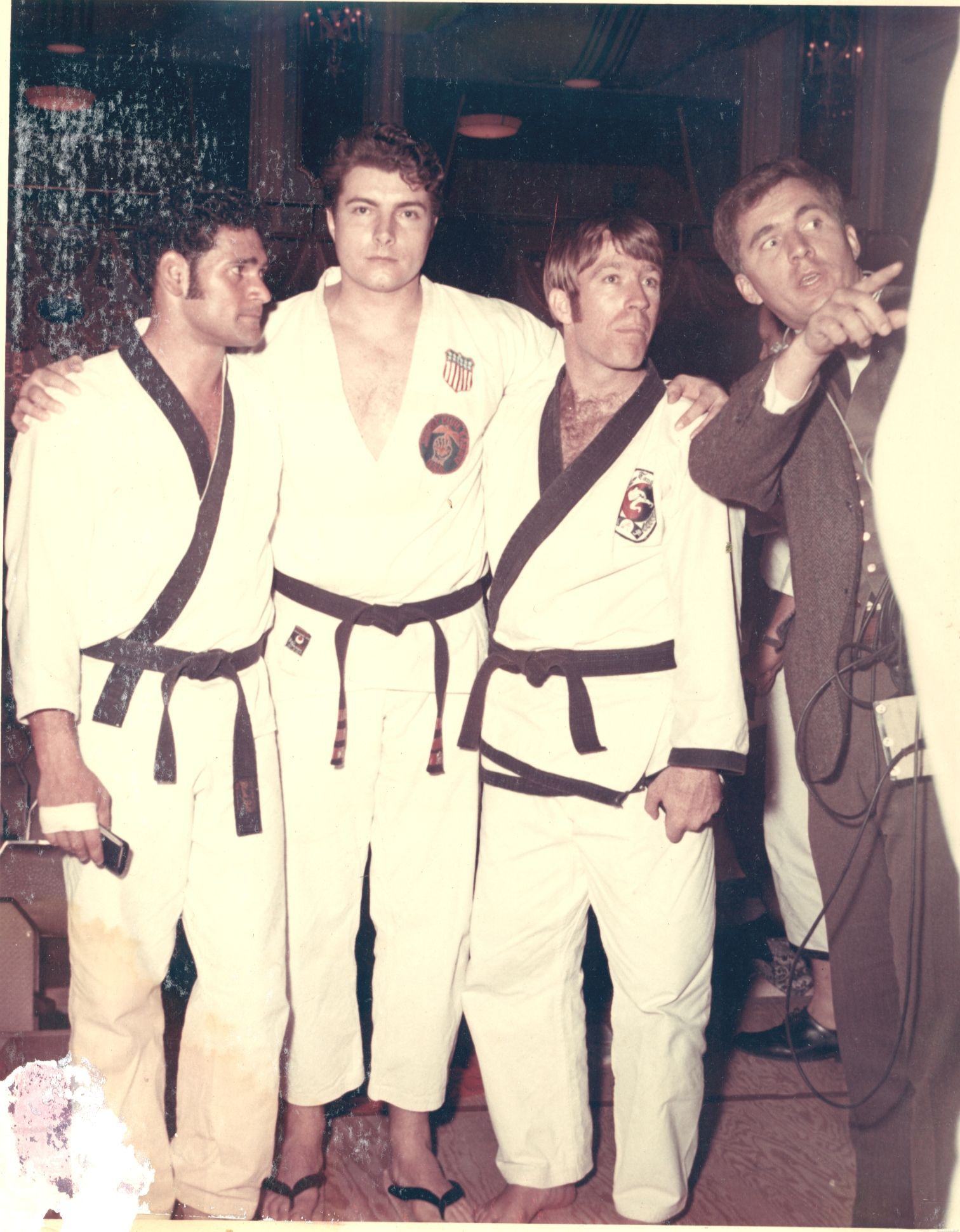 Three martial artists in white uniforms with black belts pose with a man holding a microphone, possibly at an event.