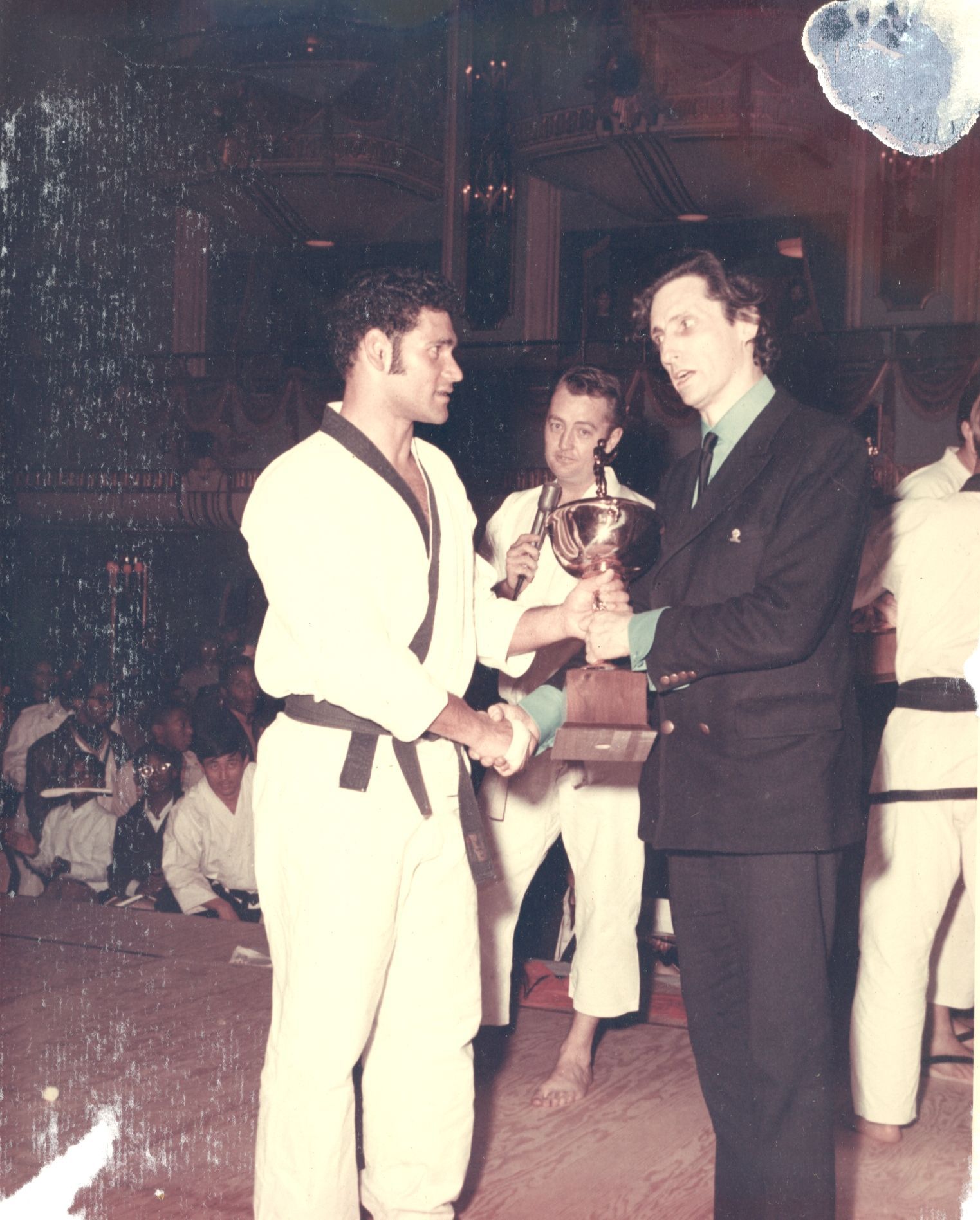 A man in a white karate uniform with a black belt receives a trophy from a man in a suit at an event. 