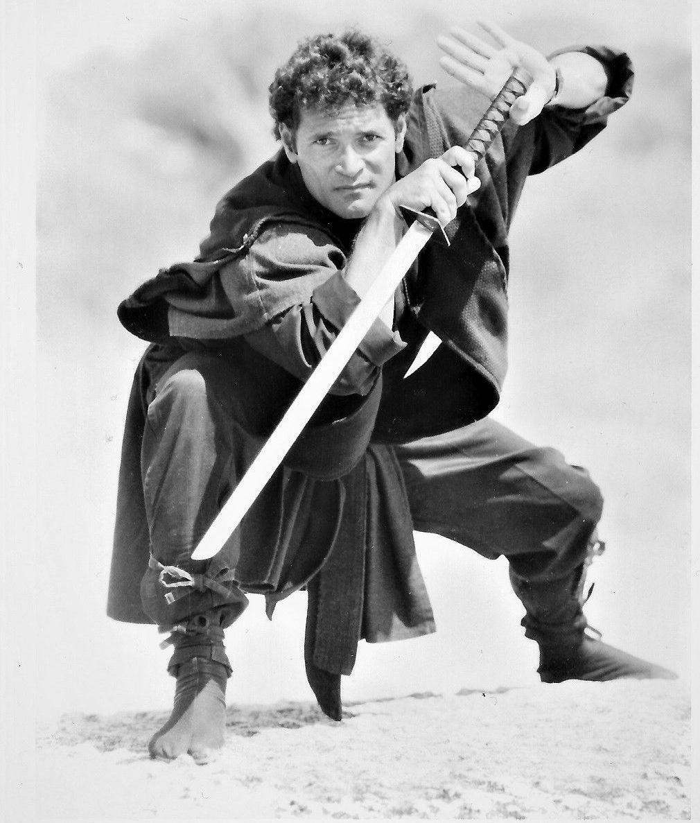 Man in ninja attire, holding a katana sword, in a fighting stance. He’s outdoors with a focused expression.