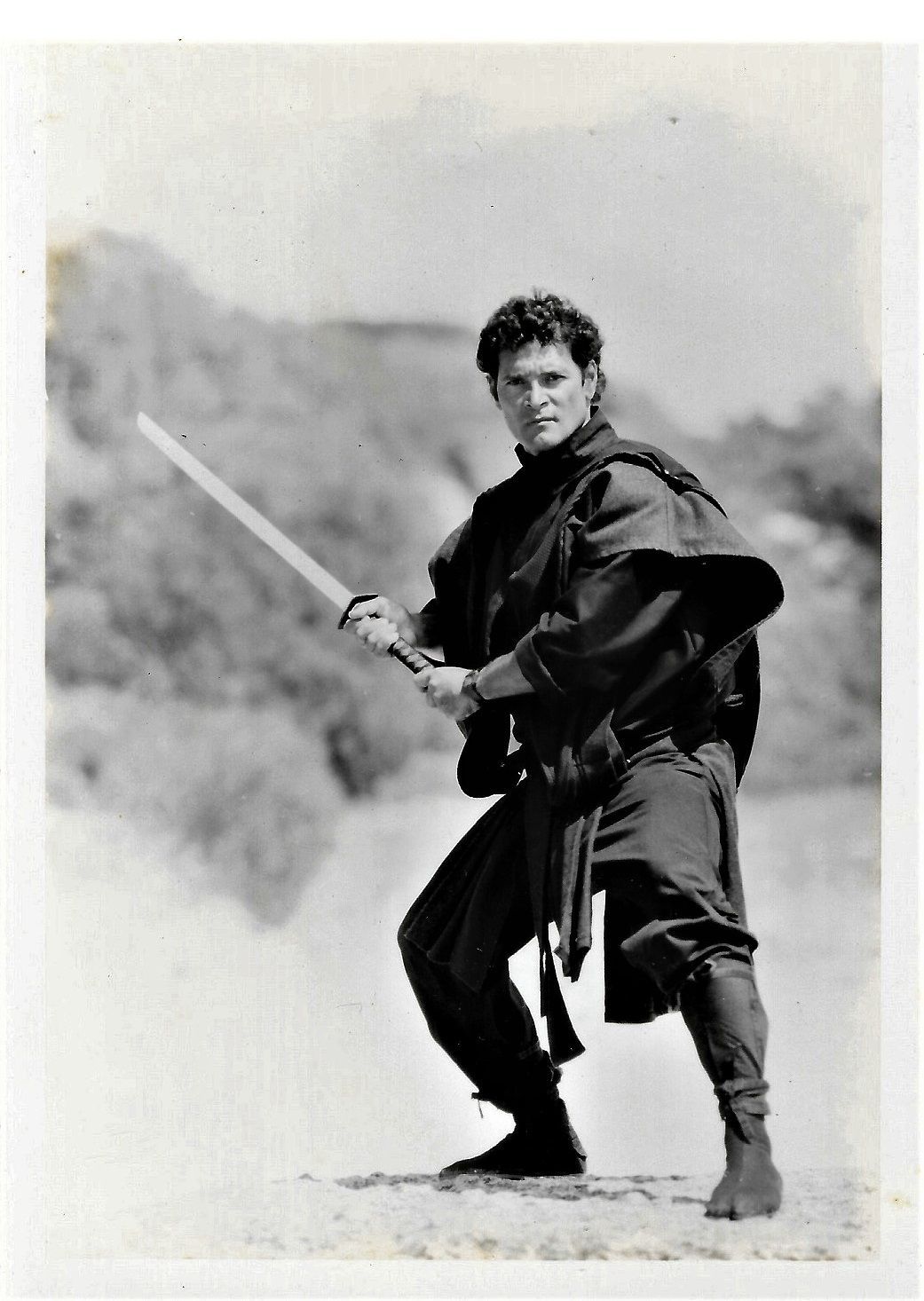 A man in a black ninja outfit holds a katana, poised in a fighting stance, outdoors.