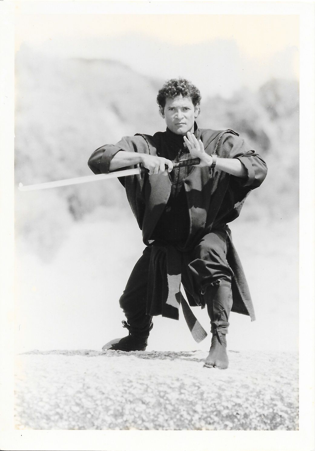 Man in martial arts pose with sword, outdoors. He wears a dark outfit, with a serious expression.