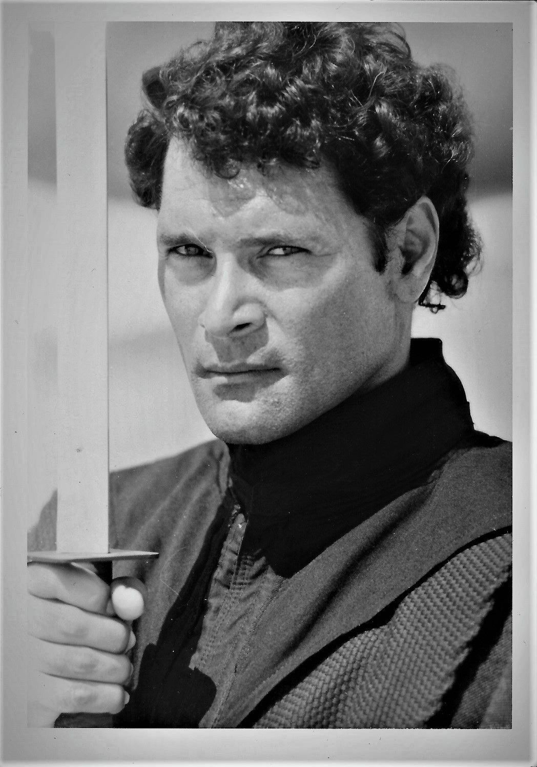 A black and white portrait of Mike holding a sword, looking directly at the camera. 