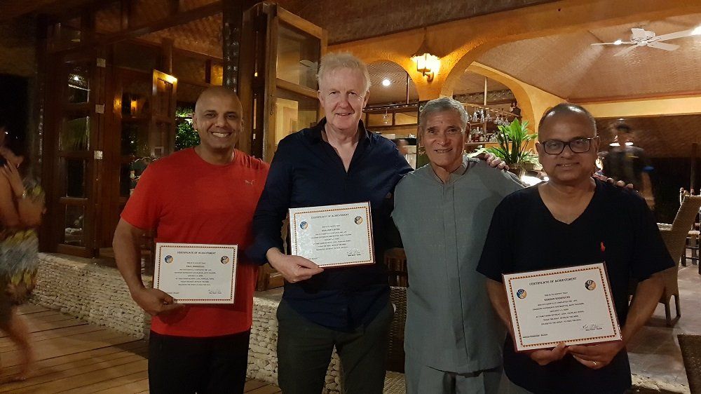 Four men holding certificates, smiling, likely after an event. They stand inside a restaurant with a warm ambiance.