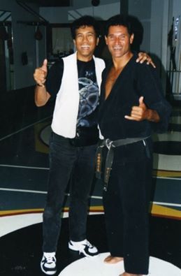 Two men posing in a gym. One in a karate uniform with a black belt, the other in a white vest giving a thumbs up.