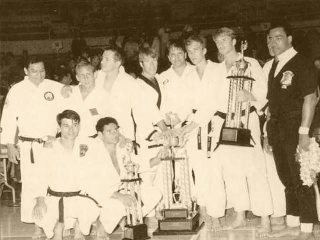 Group of karate tournament winners holding trophies, posing together on a stage. 