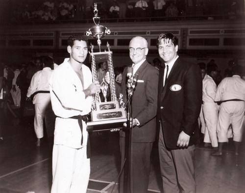 Karate champion holding a trophy, flanked by two men in suits, likely at an awards ceremony in a gym.