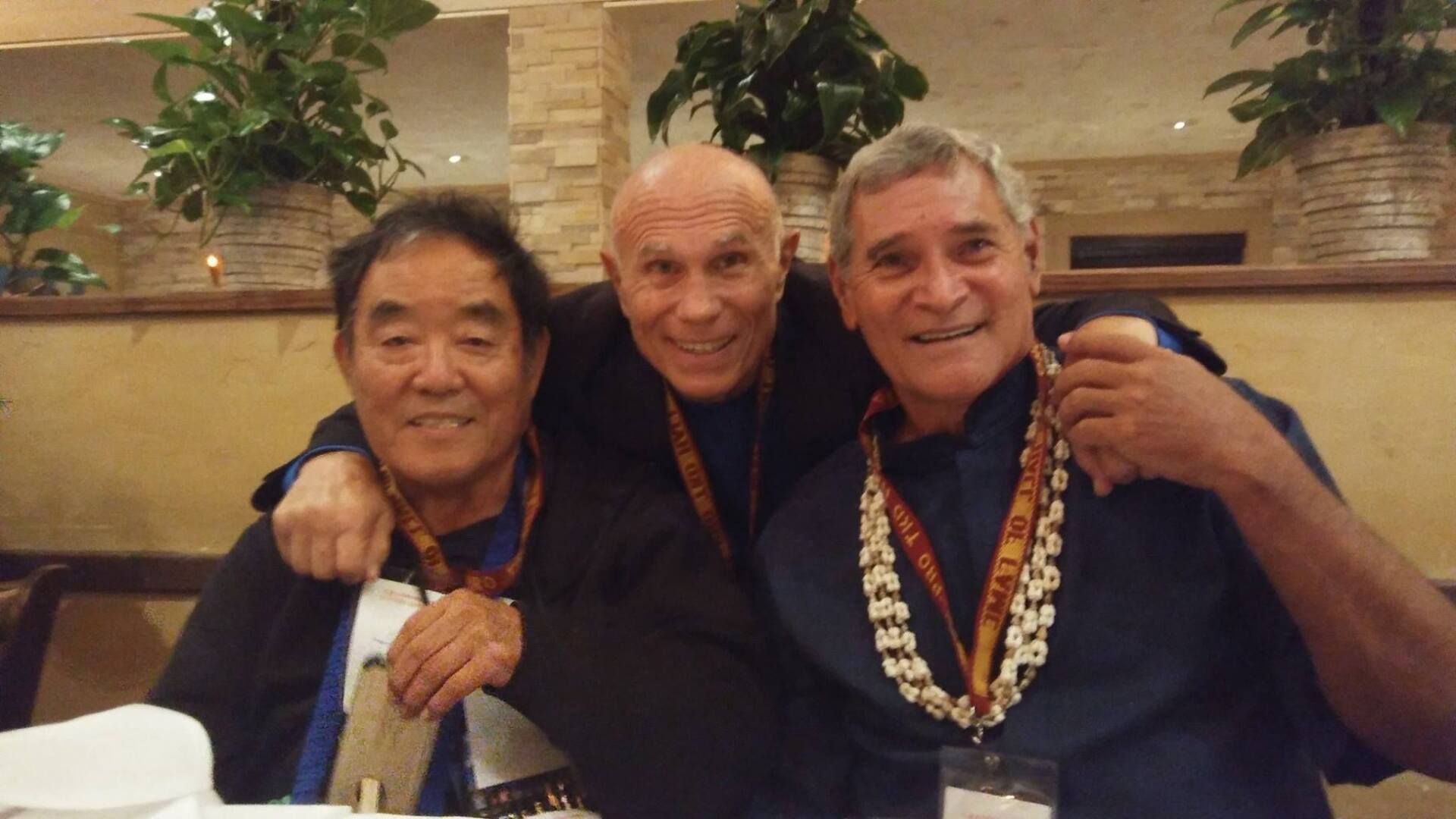 Three men in a restaurant setting smiling at the camera, one with arms around the others; all wearing leis and lanyards.