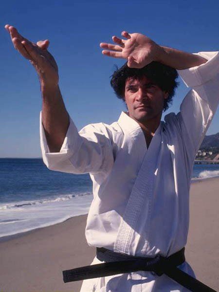 Man in white karate uniform on a beach performs a defensive hand gesture, ocean in background.