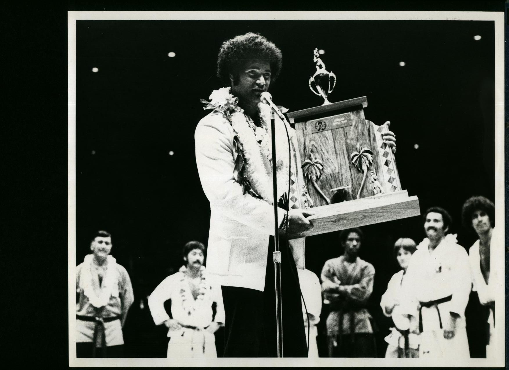 Man holding a large trophy at a podium. He wears a white jacket and holds a microphone. Other people stand behind him.