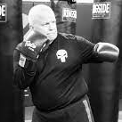 A bald man in boxing gloves throws a punch in a gym. He wears a black shirt with a skull logo.