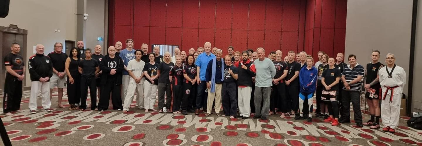 A large group of people in martial arts uniforms pose in a conference room. Red carpet and backdrop.